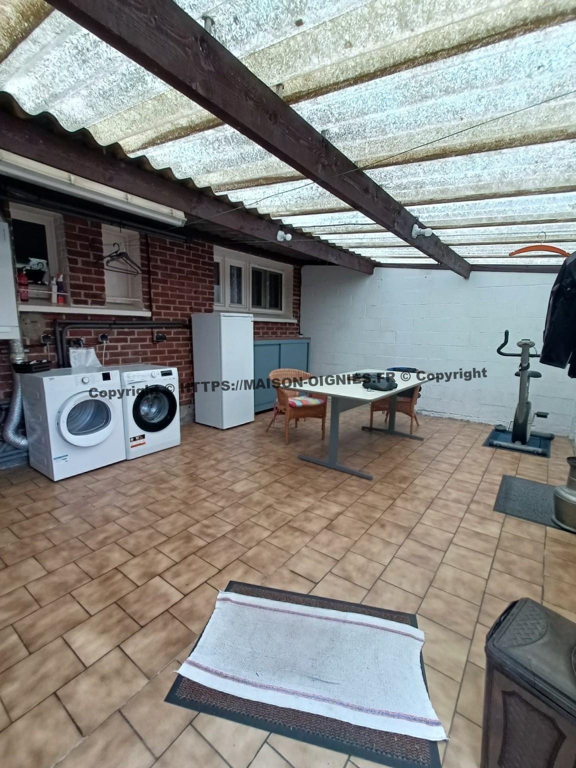 L'image montre la buanderie depuis la porte qui donne sur l'allée du jardin, une chaudière Wiessmann parfaitement entretenue est installée sur la gauche, sèche-linge, machine à laver, un congélateur et une armoire de rangement suivent vers la droite, une table y est installée pour l'hivernage des plantes ou autres utilisations