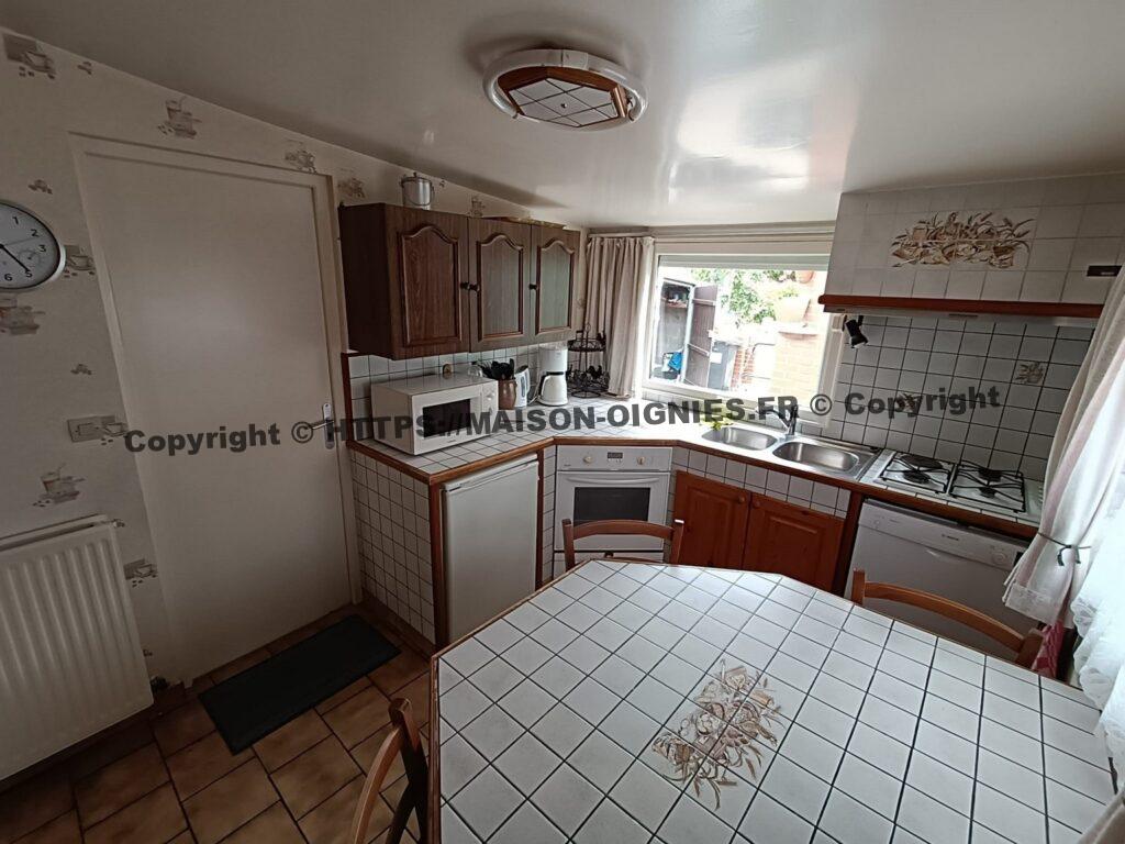 l'image, en léger surplomb, montre la cuisine avec au premier plan la table carrelée fixe, des chaises, puis un meuble où sont intégrés un four électrique, un évier double en inox, une table de cuisson gaz et électricité, un petit réfrigérateur et un lave vaisselle sont également disponible