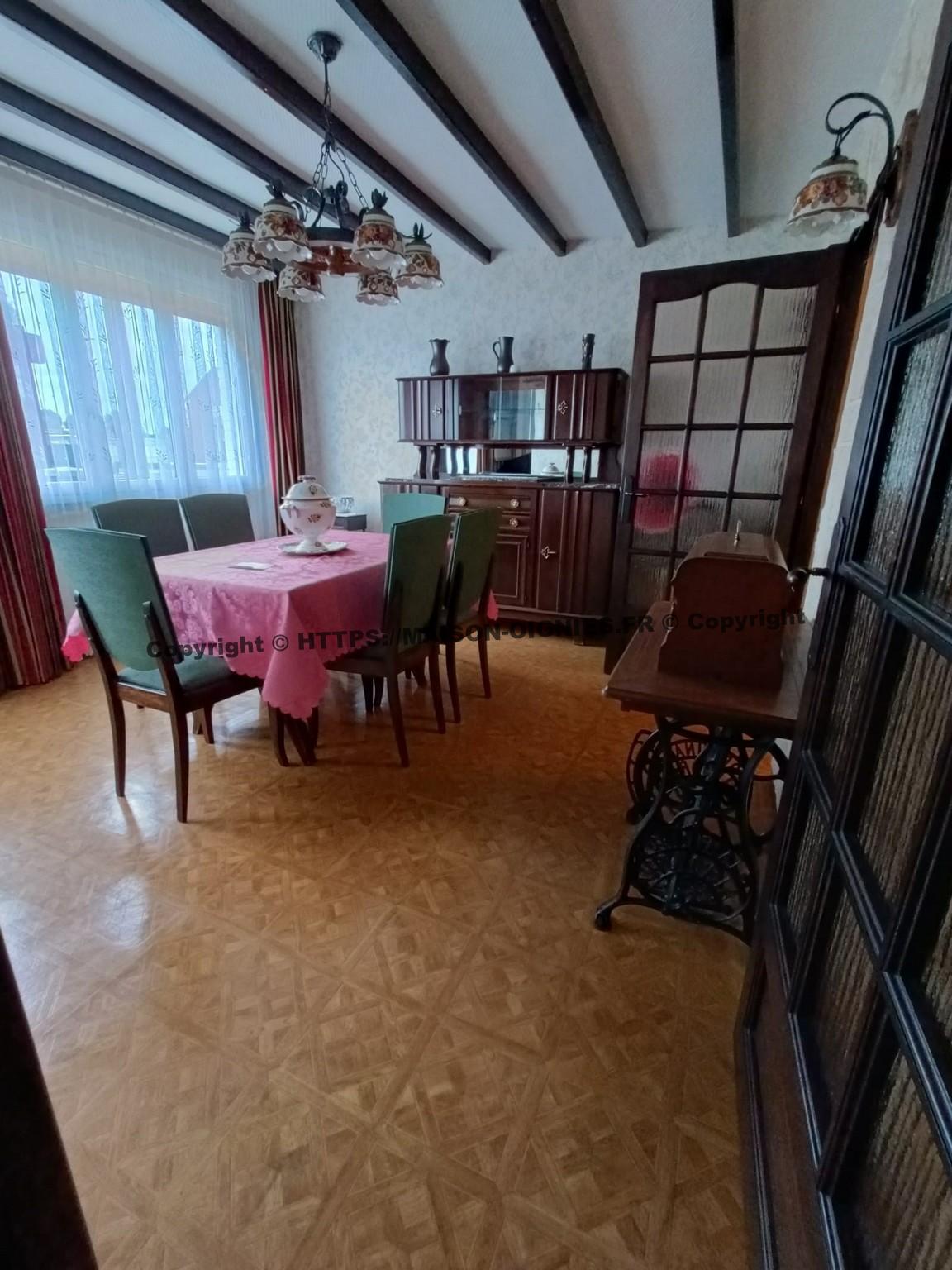 l'image montre la salle à manger depuis le couloir d'entrée, plafond blanc avec poutres lisses de décoration, le sol est synthétique représentant un parquet bois dit "versailles", une table avec six chaises au centre de style années 60, surplombé d'un lustre bois avec caches céramiques, une armoire vernie de style ancien, et une ancienne machine à coudre