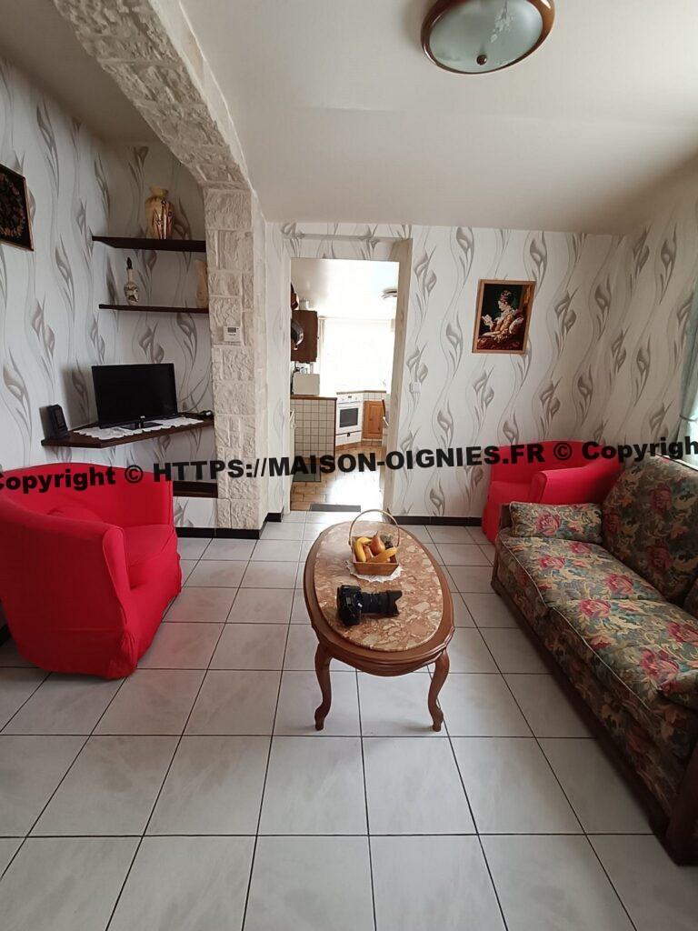 L'image montre le salon vu depuis la porte d'entrée avant et couloir, une banquette 3 places à droite suivie d'un fauteuil rouge, une table basse au milieu et le second fauteuil rouge sur la gauche devant des étagères au fond à gauche dont une large pour un téléviseur, le sol est carrelé en gris légèrement marbré au fond la porte d'entrée de la cuisine